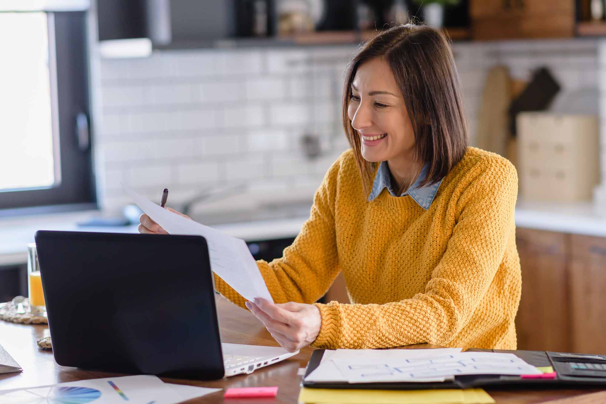 Woman sitting in her kitchen looking through papers next to her laptop