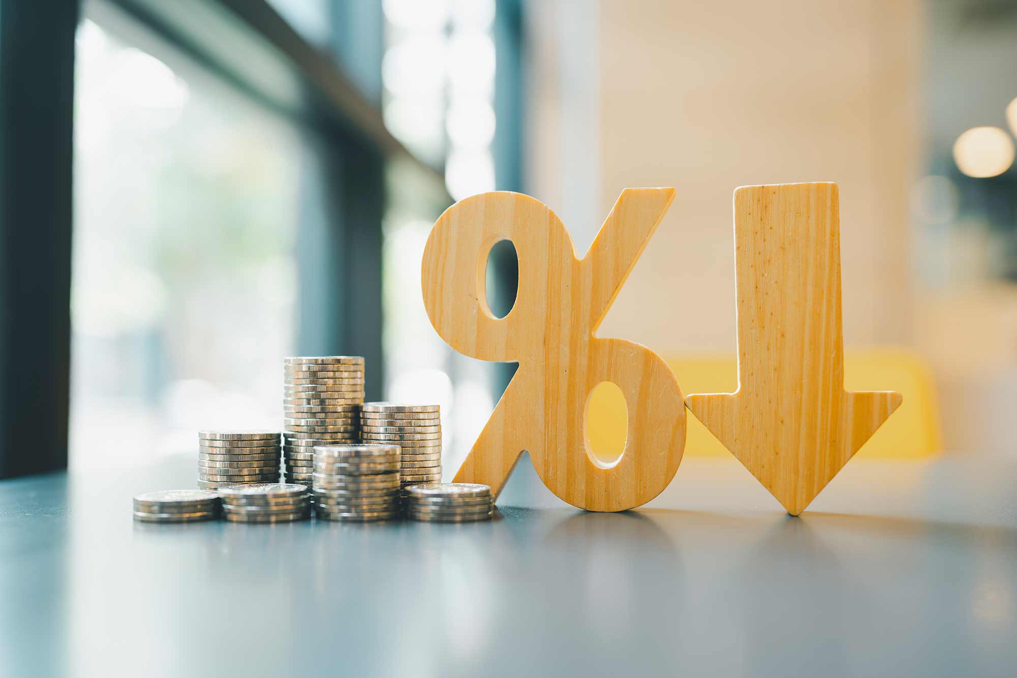 A pile of money, a percentage sign, and an arrow facing down all sitting on a table in front of a window