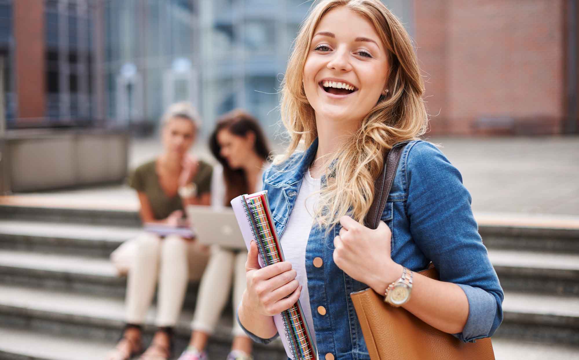 Young girl with blond hair holding school books and a bag and smiling in front of a school building