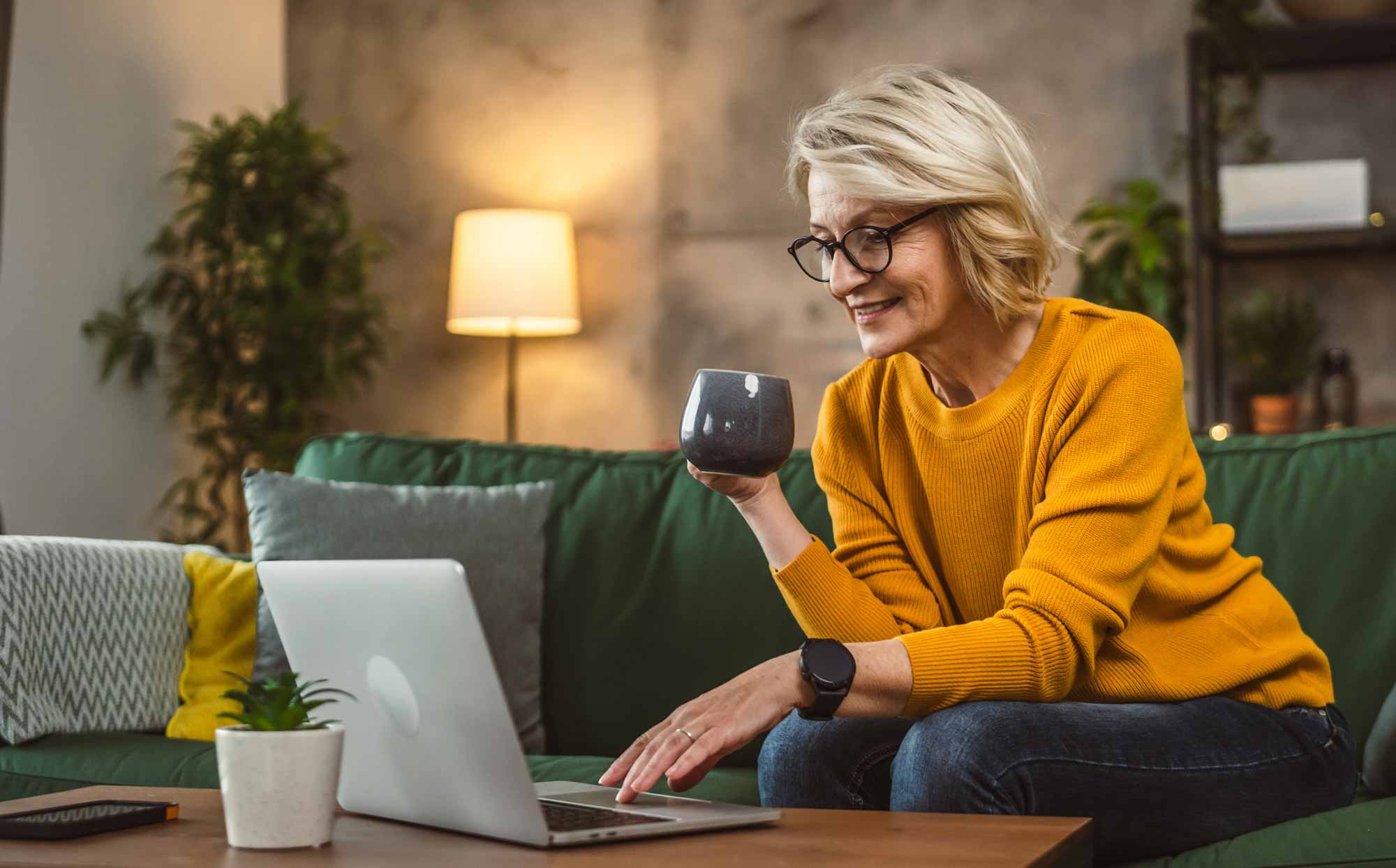 Woman sitting on her couch holding a coffee while browsing on her laptop
