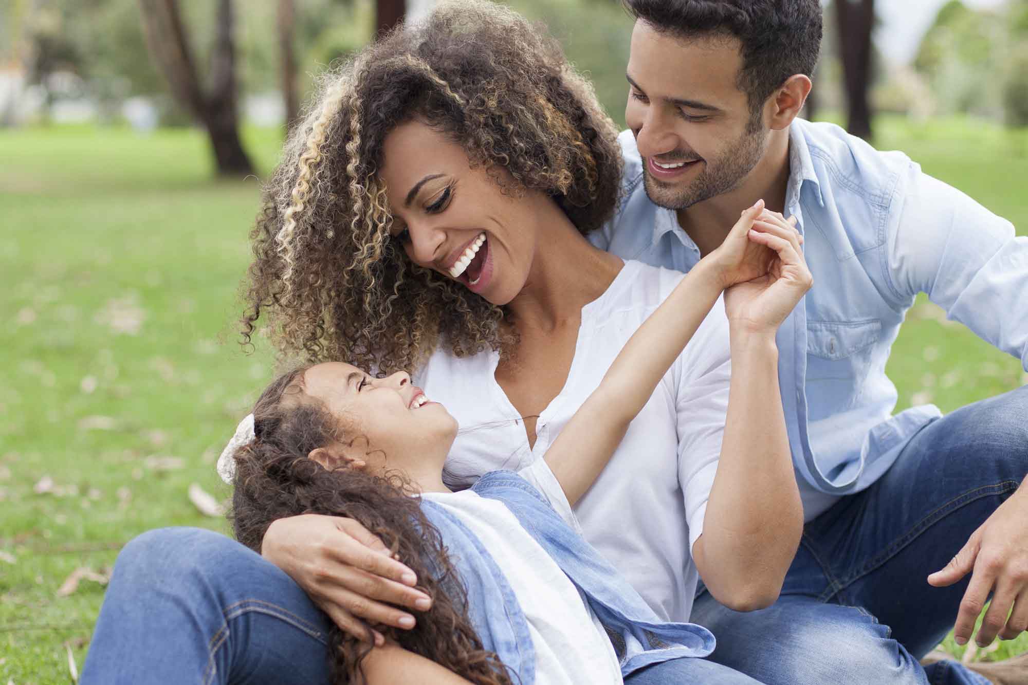 A young family sitting on the ground in a park laughing together.