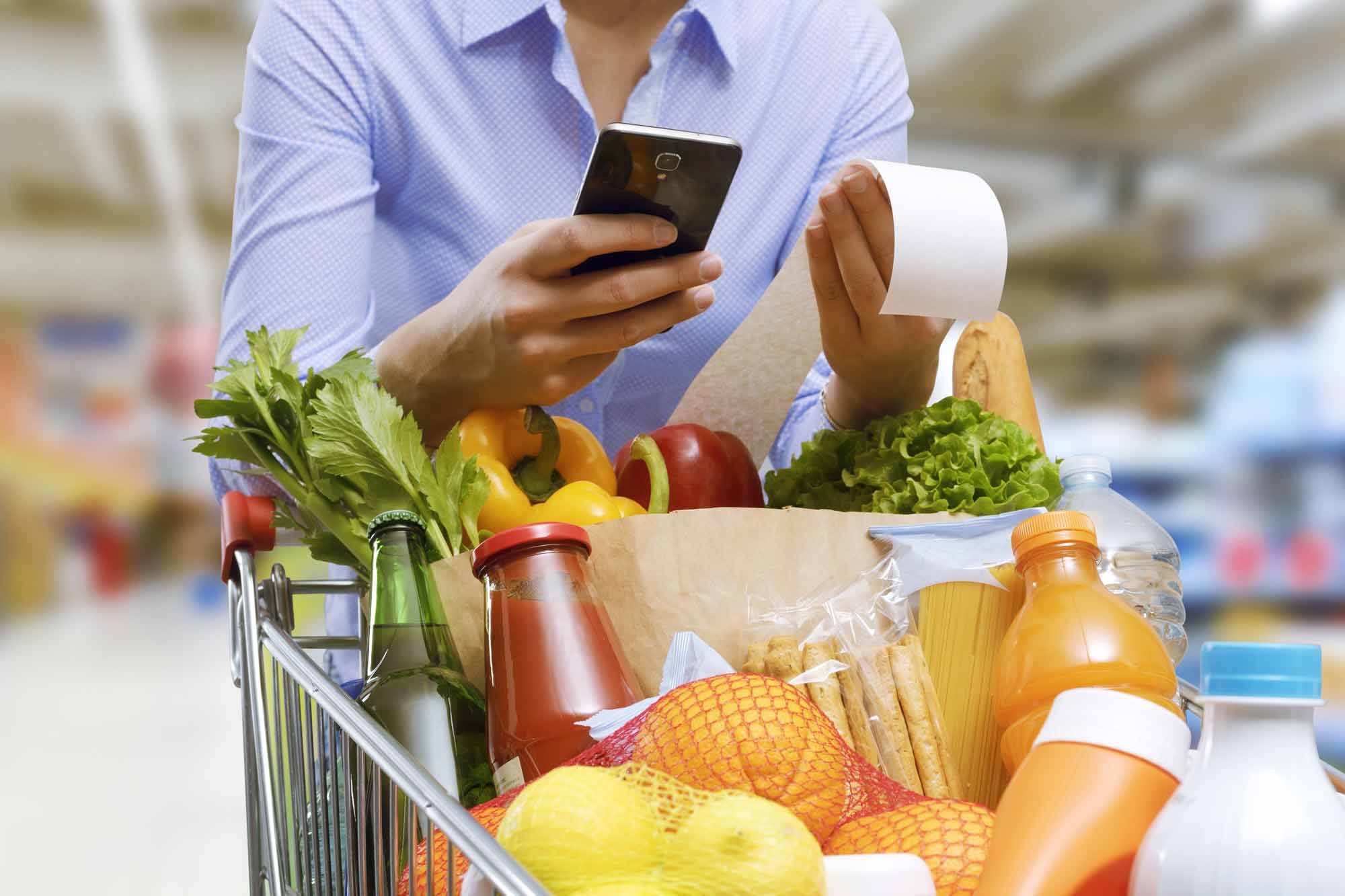 A woman pushing a cart full of groceries while looking at her receipt and the calculator on her phone