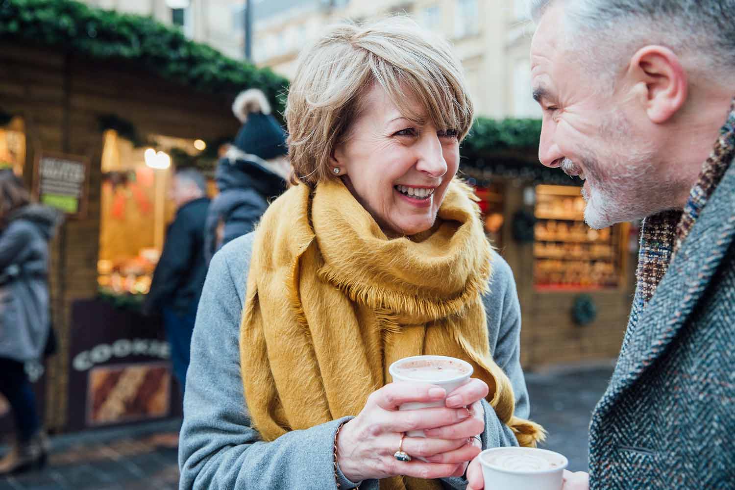 An older couple somewhere in Europe enjoying some hot chocolate together outdoors in front of a small shop.